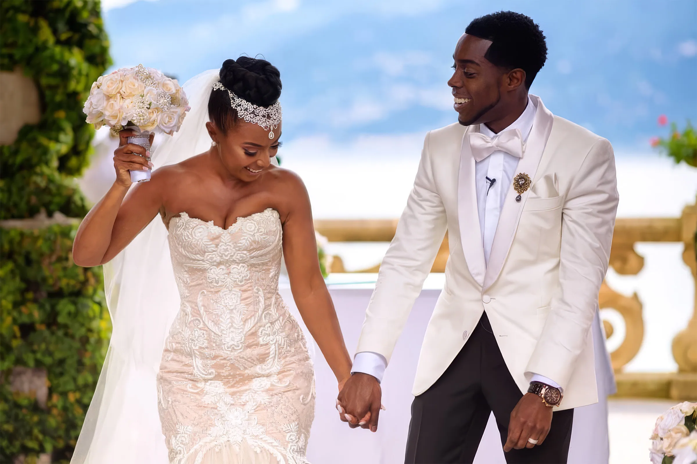 Denise and her husband walk the aisle beneath ivy-covered arches at Lake Como, Italy. She raises her bouquet in a bespoke Kosibah couture mermaid gown with a long tulle train; the lake and mountains are behind them.