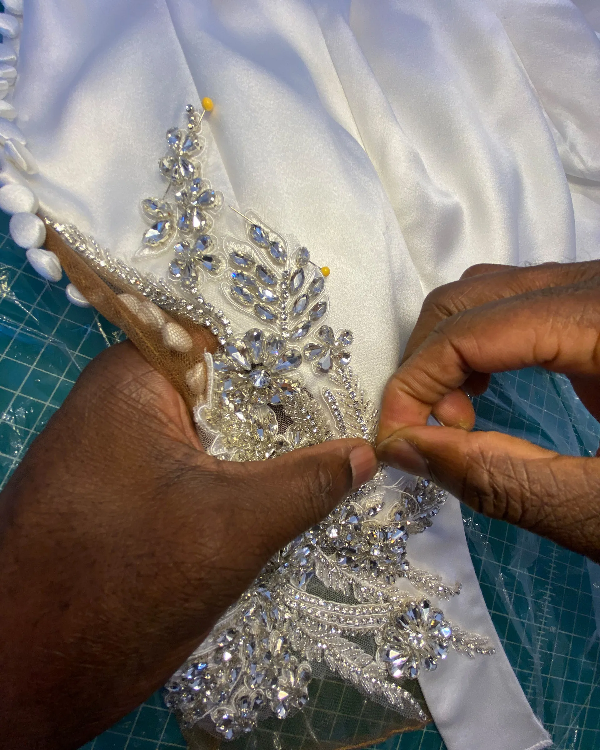 Hands sewing a crystal-beaded appliqué onto an ivory satin gown, the appliqué held in place with pins.